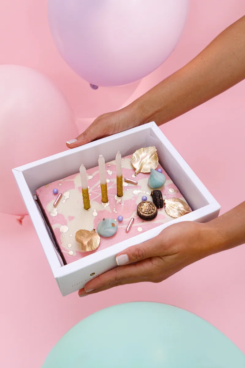 Manos sosteniendo una caja con torta decorada y velas de Boreal, entre globos rosados